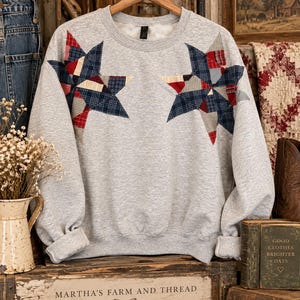 May include: A light gray crewneck sweatshirt featuring two large, appliqued stars on the chest. The stars are constructed from a patchwork of various fabric scraps in shades of navy blue, red, and cream, showcasing a mix of plaid and solid patterns.