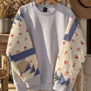 May include: A light gray crewneck sweatshirt featuring long sleeves with a quilted patchwork design. The sleeves incorporate cream fabric with a small red and yellow floral print, accented by solid blue and dark patterned geometric fabric panels.