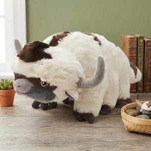 May include: A plush toy of a six-legged creature with cream-colored fur, dark brown markings on the head and back, curved gray horns, and a gray snout. The creature has large dark eyes and a wide, flat tail. A smaller version is in a basket.