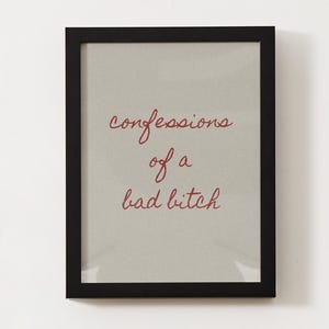 May include: A rectangular wall art piece featuring a black frame and a light gray background. The text "confessions of a bad bitch" is written in the center in a red, cursive, handwritten-style font. The design is simple and centered.