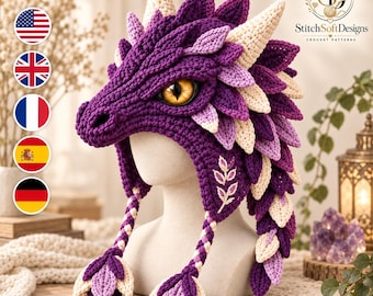 May include: A crochet dragon hat in shades of purple, lavender, and cream, featuring a textured scale pattern, a prominent snout, and a single large yellow eye with a black slit pupil. Two braided earflaps with leaf-shaped ends hang down.