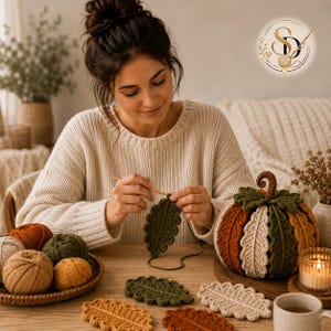 Female crochet artisan working on an olive green oak-leaf coaster, with matching finished leaves and the assembled crochet pumpkin displayed nearby.