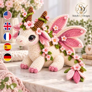 May include: A cream-colored crochet dragon plush toy featuring large dark eyes, brown horns, and pink-tipped wings. The creature is adorned with green vines and small pink cherry blossom flowers that wrap around its head, body, and long tail.