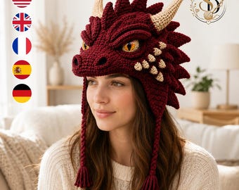 May include: A deep red crochet dragon hat featuring textured scales, two beige horns, and realistic yellow eyes. The hat includes ear flaps with braided tassels hanging down on each side. The design mimics a dragon head with a prominent snout and crest.