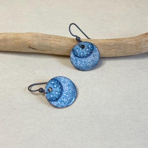 May include: A pair of round, blue and white enamel earrings with a speckled pattern. The earrings have dark gray ear wires and a small bead detail. The earrings are displayed on a light beige surface.