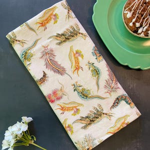 May include: A folded cream-colored kitchen towel with a colorful sea slug pattern. The towel is next to a green plate with a muffin and a sprig of white flowers on a dark surface.