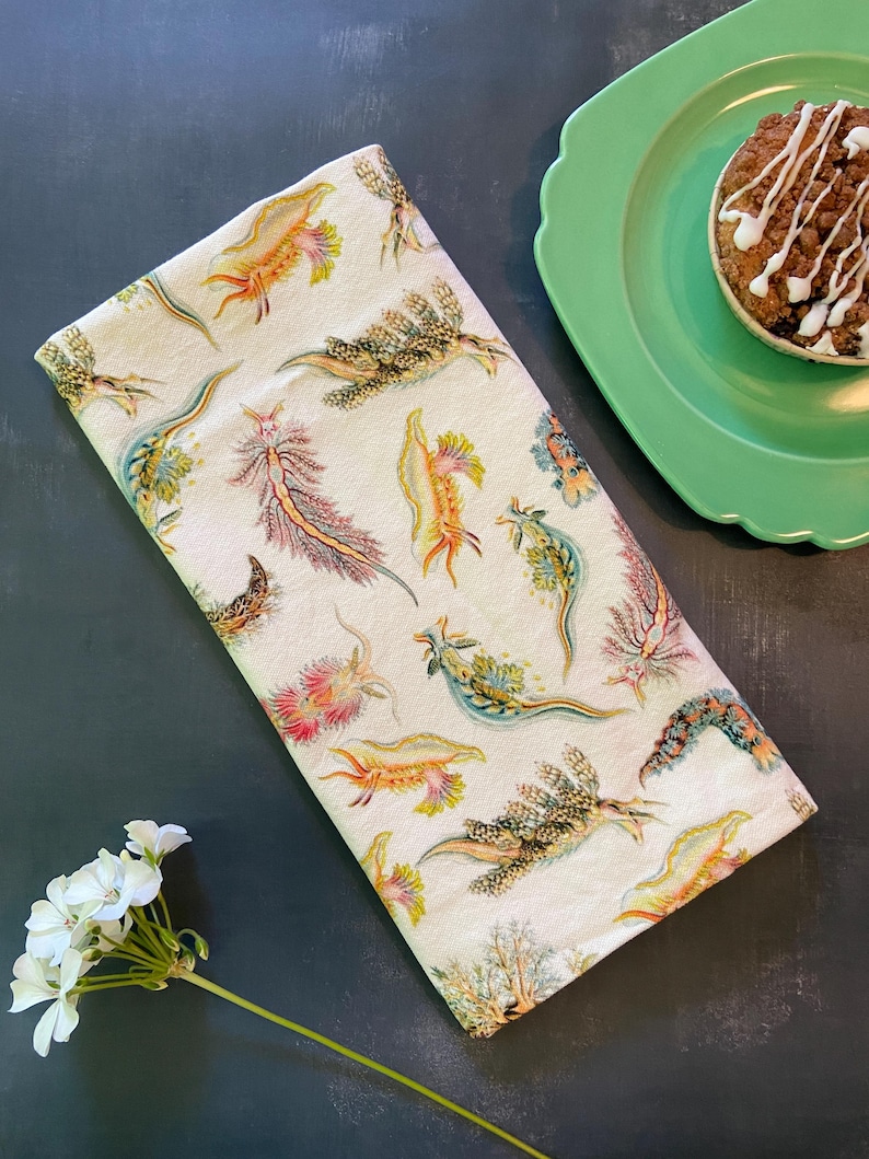 May include: A folded cream-colored kitchen towel with a colorful sea slug pattern. The towel is next to a green plate with a muffin and a sprig of white flowers on a dark surface.