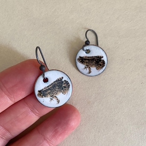 May include: A pair of silver dangle earrings with white ceramic round charms featuring a black and brown frog illustration.