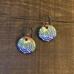 May include: A pair of earrings with a colorful, circular design. The earrings are made of metal and have a black wire hook. The design features a white, circular pattern on a yellow, orange, blue, and purple background.