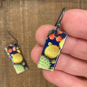 May include: Rectangular earrings with a dark blue background and a pattern of yellow, orange, and green circular shapes. The earrings have a silver-colored hook and are held in a hand.