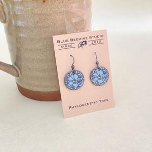 May include: A pair of silver dangle earrings with a light blue enamel disc featuring a white phylogenetic tree design. The earrings are displayed on a card with the text "BLUE BEEHIVE STUDIO SINCE 2012" and "PHYLOGENETIC TREE".
