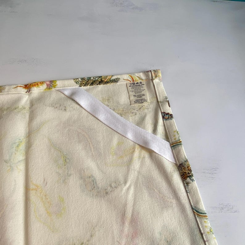 May include: Cream-colored kitchen towel with a colorful floral pattern. The towel has a white ribbon loop for hanging and a tag with care instructions. The floral design includes yellow, green, and blue accents.