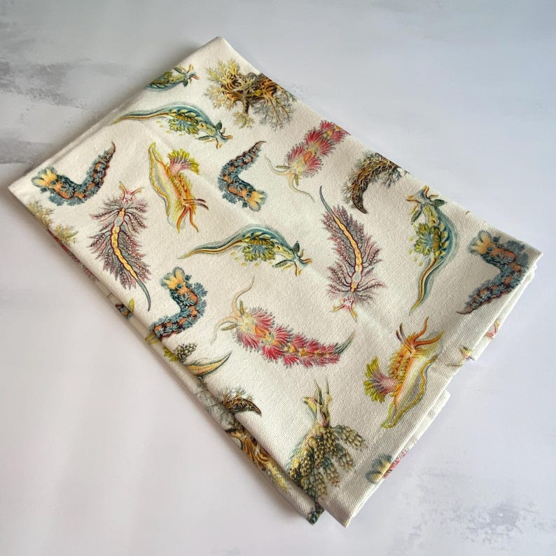 May include: Folded white kitchen towel featuring a repeating pattern of colorful sea slugs in shades of yellow, pink, green, and blue. The sea slugs are illustrated in a detailed, artistic style. The towel is made of a soft, absorbent fabric.