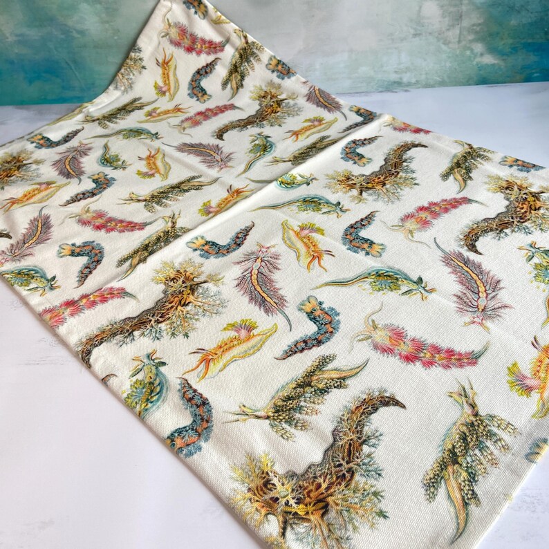 May include: Cream-colored fabric with a repeating pattern of colorful sea slugs in shades of brown, green, pink, and yellow. The sea slugs are in various shapes and sizes, creating a unique and artistic design. The fabric is folded, showing the pattern's continuity.