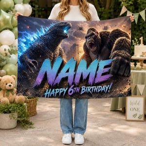 Op de afbeelding: Een rechthoekige banner met een grafische illustratie van Godzilla en King Kong die tegenover elkaar staan tegen een stormachtige lucht en stadsachtergrond. De tekst NAME en HAPPY 6TH BIRTHDAY staat in grote, dikke blauw-paarse letters.