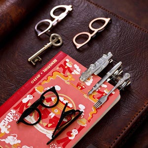 Puede incluir: Una colección de accesorios metálicos que incluye dos pares de clips con forma de gafas, una llave de estilo antiguo, un clip con forma de coche, un clip con forma de bolígrafo y un clip con forma de tenedor sobre un cuaderno rojo ilustrado.