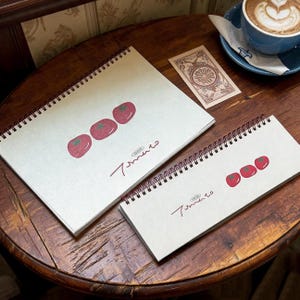 May include: Two spiral-bound notebooks with off-white covers, each featuring a graphic of three red tomatoes and the handwritten text "Tomato". The larger notebook is rectangular, while the smaller one is long and narrow. A small rectangular card with a circular design and a blue cup of coffee are also present.