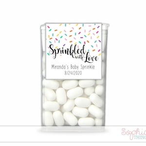 May include: A clear plastic container filled with white mints. A label on the front reads "Sprinkled with Love" with a colorful sprinkle pattern. The label also includes the text "Miranda's Baby Sprinkle 8/24/2020".