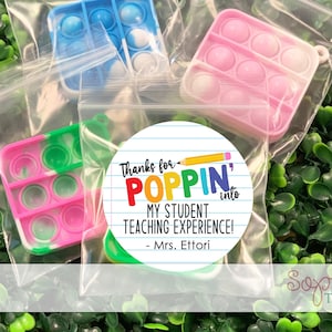 May include: A round sticker with a white background and black lines. The sticker says "Thanks for POPPIN' into MY STUDENT TEACHING EXPERIENCE! - Mrs. Ettori". The sticker has a rainbow colored pencil on it. There are three colorful pop-it fidget toys in the background.