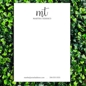 May include: A white notepad with the initials "mt" in elegant black script at the top, followed by the name "Martha Tedesco." The bottom of the notepad includes an email address and phone number in a simple font. The background is a dense arrangement of green leaves.