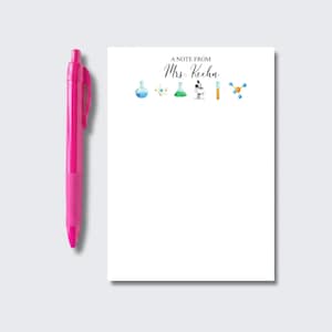 May include: A bright pink retractable ballpoint pen next to a white notepad featuring the text A NOTE FROM Mrs. Keehn at the top, accompanied by a row of colorful watercolor science illustrations including flasks, a microscope, and molecules.