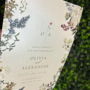 May include: A rectangular wedding invitation card on cream-colored paper featuring a botanical border of white flowers, green leaves, and berries. The text reads: O A, KINDLY JOIN US FOR THE WEDDING OF OLIVIA and ALEXANDER, AUGUST 20TH AT 4 PM, 123 ANYWHERE ST.