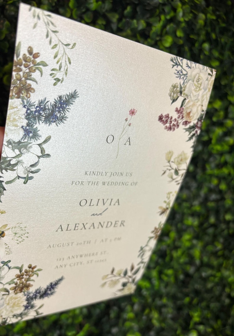 May include: A rectangular wedding invitation card on cream-colored paper featuring a botanical border of white flowers, green leaves, and berries. The text reads: O A, KINDLY JOIN US FOR THE WEDDING OF OLIVIA and ALEXANDER, AUGUST 20TH AT 4 PM, 123 ANYWHERE ST.