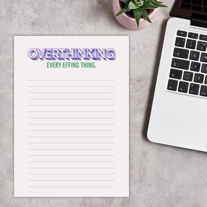 May include: A lined notepad with the text "OVERTHINKING EVERY EFFING THING" in purple and green. A laptop and a small potted plant are visible in the background. The notepad is on a gray surface.