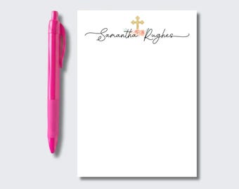 May include: A bright pink retractable ballpoint pen positioned next to a white rectangular notepad. The notepad features the name Samantha Rughes in black cursive script, topped with a gold cross icon and small pink floral accents.