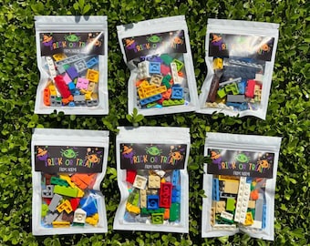May include: Six clear plastic pouches filled with assorted colorful plastic building bricks. Each pouch features a black label at the top with the text "BRICK OR TREAT" and "FROM NOEMI" accompanied by illustrations of a green spider and orange hats.