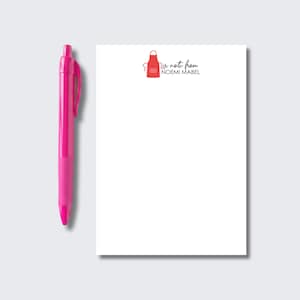 May include: A bright pink retractable ballpoint pen positioned next to a rectangular white notepad. The notepad features a small red apron icon at the top center, followed by the black printed text, a note from NOEMI MABEL.