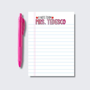 May include: A bright pink retractable ballpoint pen positioned next to a white notepad with blue horizontal lines and a vertical red margin. The top of the notepad features the printed text A NOTE FROM MRS. TEDESCO with two small red apple icons.