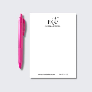 May include: A bright pink retractable ballpoint pen positioned next to a white rectangular notepad. The notepad features the black printed initials mt, the name MARTHA TEDESCO, and contact information including an email and phone number.