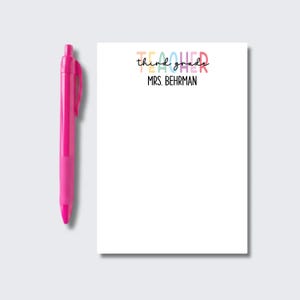 May include: A bright pink retractable ballpoint pen positioned next to a white rectangular notepad. The notepad features the colorful text "third grade" in a playful font, with the name "MRS. BEHRMAN" printed in black capital letters below.