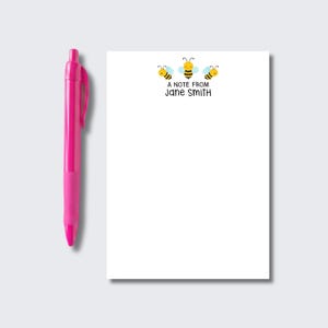 May include: A bright pink retractable ballpoint pen positioned next to a white rectangular notepad. The notepad features a graphic of three yellow and black bees at the top, with the printed text A NOTE FROM JANE SMITH centered below them.