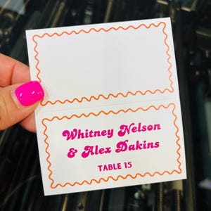 May include: A white folded place card featuring an orange scalloped border. The bottom section displays the pink text Whitney Nelson & Alex Dakins and TABLE 15. The top section is blank, providing space for additional information or guest names.