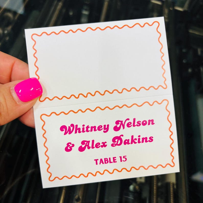 May include: A white folded place card featuring an orange scalloped border. The bottom section displays the pink text Whitney Nelson & Alex Dakins and TABLE 15. The top section is blank, providing space for additional information or guest names.