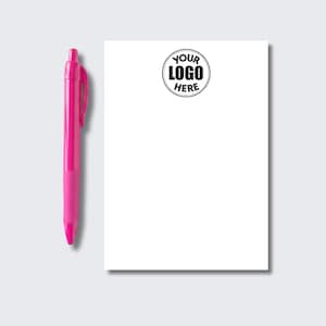May include: A bright pink retractable ballpoint pen positioned next to a rectangular white notepad. The notepad features a circular black graphic at the top center containing the text YOUR LOGO HERE in a bold, sans-serif typeface.
