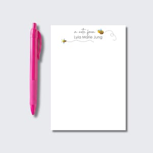 May include: A bright pink retractable ballpoint pen positioned next to a white rectangular notepad. The notepad features the printed text 'a note from Lyla Marie Jung' in black script and print, accompanied by two small yellow and black bees.