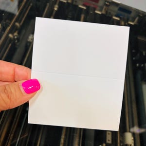 May include: A square, blank white card with a horizontal crease across the center, designed to be folded in half. The card features a smooth, matte surface and clean, straight edges, providing a simple and versatile base for custom printing.