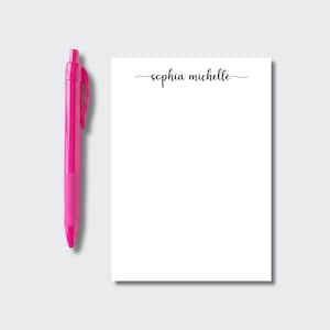 May include: A bright pink retractable ballpoint pen positioned next to a rectangular white notepad. The notepad features the name Sophia Michelle printed in a black cursive script font at the top center of the page, leaving the rest blank.