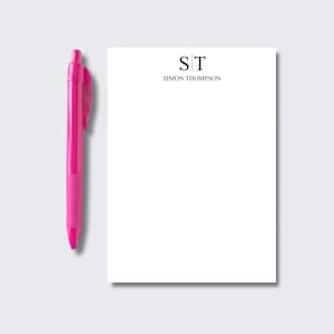 May include: A bright pink retractable ballpoint pen positioned next to a rectangular white notepad. The notepad features the black printed monogram ST at the top, followed by the name SIMON THOMPSON in a clean, black serif typeface.