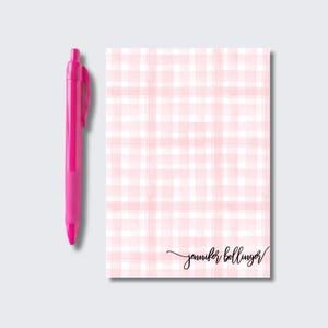 May include: A rectangular notepad featuring a light pink and white watercolor-style plaid pattern with the name jennifer bollinger written in black cursive script at the bottom right, accompanied by a solid bright pink retractable ballpoint pen.