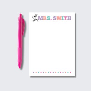 May include: A white rectangular notepad featuring the text 'a note from MRS. SMITH' in multicolored block letters at the top and a row of small colored dots at the bottom, accompanied by a bright pink retractable ballpoint pen positioned to the left.