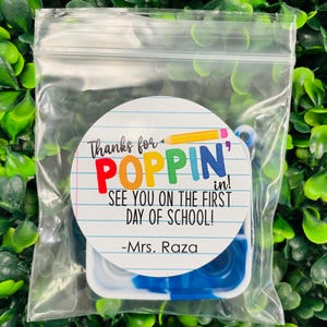 Puede incluir: Una pegatina circular blanca con el texto "Thanks for POPPIN' in! See you on the first day of school! -Mrs. Raza" en letras de colores. La pegatina est&aacute; en una bolsa de pl&aacute;stico transparente con un juguete fidget azul y blanco.