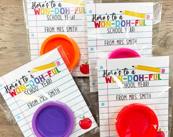 Może przedstawiać: Cztery przezroczyste plastikowe torebki, każda zawierająca małą kartkę z tekstem "Here's to a WON-DOH-FUL SCHOOL YEAR!" i "FROM: MRS. SMITH" oraz mały pojemnik z plasteliną. Plastelina jest w różnych kolorach: pomarańczowym, różowym, fioletowym i czerwonym.