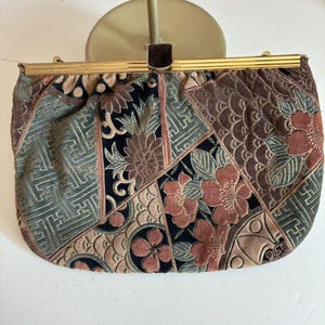 May include: A rectangular clutch bag featuring a multi-panel fabric design with a mix of floral, geometric, and wave patterns in shades of muted red, sage green, black, and tan. The bag has a gold-toned metal frame and a dark brown rectangular clasp.