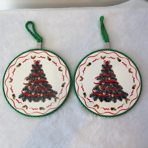 May include: Two circular white ceramic ornaments featuring a central green Christmas tree illustration decorated with red ribbons and gold ornaments. Each piece is framed by a green braided rope border and includes a matching green hanging loop.
