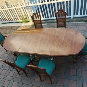 May include: An oval wooden dining table with a smooth, light brown finish and a visible grain pattern, accompanied by six chairs. The chairs feature dark wood frames, woven cane backrests, and seat cushions upholstered in a solid teal fabric.