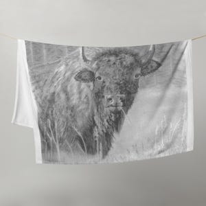 May include: A soft throw blanket featuring a detailed black and white pencil sketch of a bison standing in a field of tall grass. The illustration captures the animal's thick fur, curved horns, and focused gaze, rendered in shades of gray.
